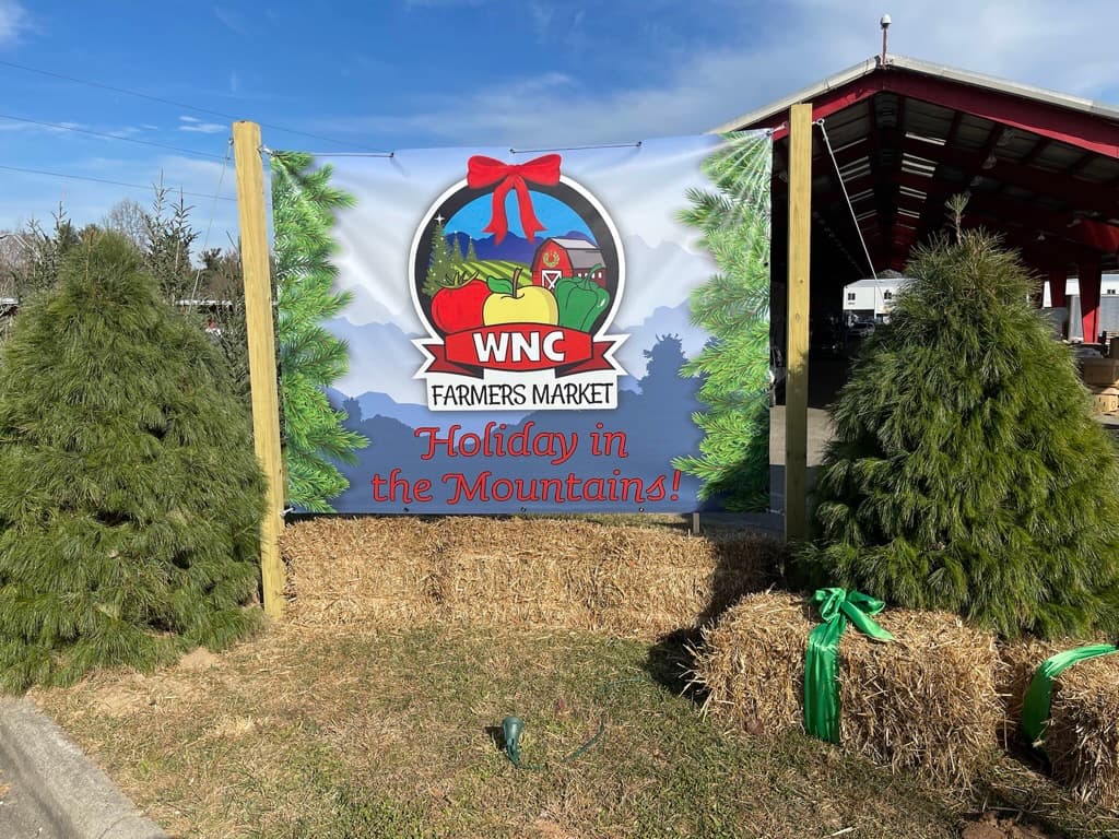 A Western North Carolina Farmers Market Holiday in the Mountains banner among Christmas trees